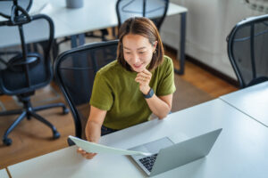 Smiling asian woman working on a laptop and reviewing a document at her office desk, highlighting her focus and happy professional engagement in a modern workspace
